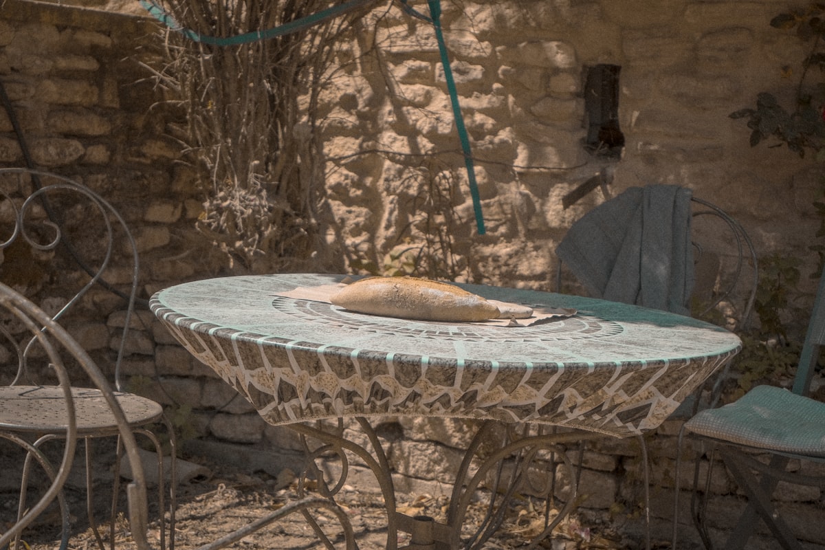 A charming outdoor table with a colorful mosaic top is set with a fresh loaf of bread, inviting relaxation in a sun-dappled courtyard. Surrounded by wrought iron chairs and stone walls, this cozy corner reflects the warm, welcoming atmosphere of a Provencal farmhouse related to Add a Touch of French Country Charm with the Perfect Valance