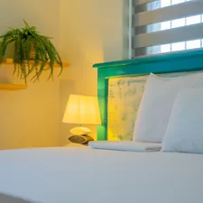 A cozy and minimal bedroom corner featuring a white bed with soft pillows, a rustic teal headboard, a warm bedside lamp, and a hanging green fern. The soft lighting and natural accents create a calming, restful ambiance. related to Chill Out with Light Blue Bedding