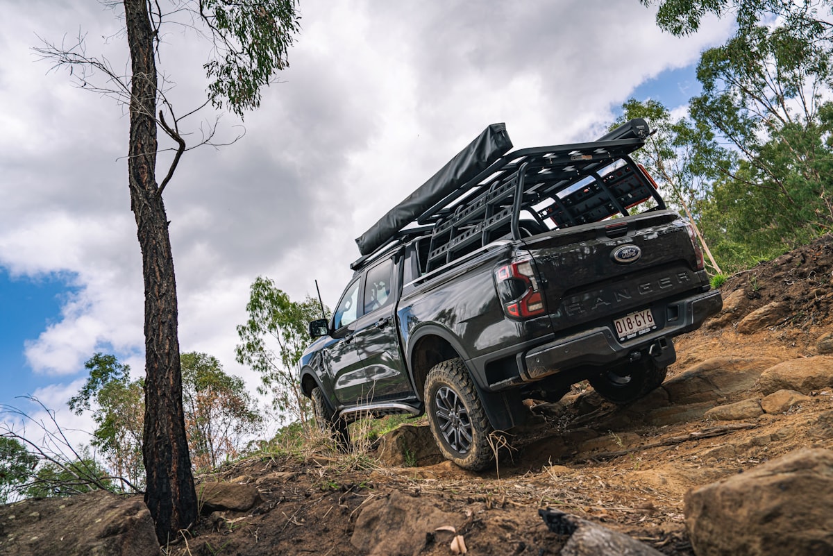 A dark grey Ford Ranger Wildtrak climbs a steep, rocky technical section of a trail. The vehicle is fully outfitted for long-range overlanding with a heavy-duty bed rack, side awning, and mud-terrain tires. The low-angle perspective emphasizes the suspension work and rugged terrain of the Australian outback. related to Ford Ranger Bed Cover: Essentials to Know