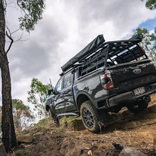A dark grey Ford Ranger Wildtrak climbs a steep, rocky technical section of a trail. The vehicle is fully outfitted for long-range overlanding with a heavy-duty bed rack, side awning, and mud-terrain tires. The low-angle perspective emphasizes the suspension work and rugged terrain of the Australian outback. related to Ford Ranger Bed Cover: Essentials to Know