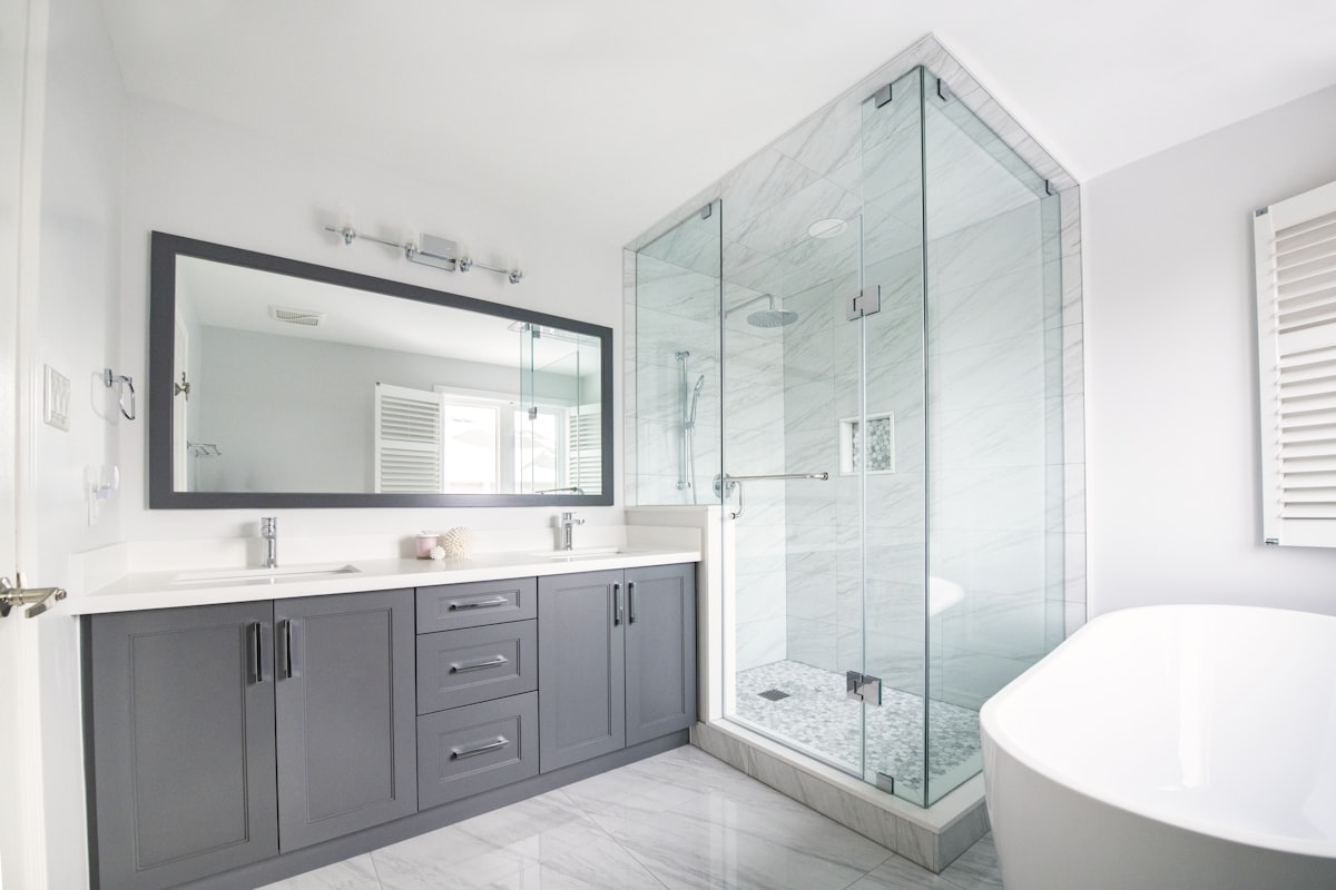 A Modern Bathroom with a bright and airy design. The space includes a spacious glass-enclosed shower with modern chrome fixtures and marble-patterned tiles, creating a luxurious feel. A sleek double-sink vanity with gray cabinetry and a large, framed mirror adds both functionality and style. The room is finished with glossy marble-look flooring and a freestanding white bathtub, while natural light streams in through white shuttered windows, enhancing the clean and sophisticated atmosphere. Get a free quote today @https://smartreno4u.ca/free-quote/ related to Bathroom Valances Ideas