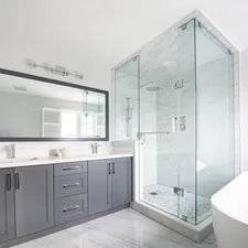 A Modern Bathroom with a bright and airy design. The space includes a spacious glass-enclosed shower with modern chrome fixtures and marble-patterned tiles, creating a luxurious feel. A sleek double-sink vanity with gray cabinetry and a large, framed mirror adds both functionality and style. The room is finished with glossy marble-look flooring and a freestanding white bathtub, while natural light streams in through white shuttered windows, enhancing the clean and sophisticated atmosphere. Get a free quote today @https://smartreno4u.ca/free-quote/ related to Bathroom Valances Ideas