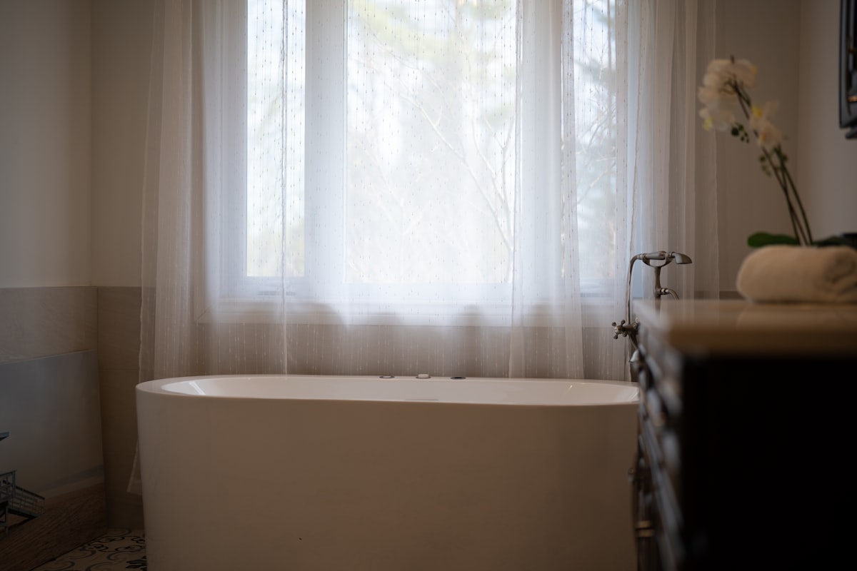 A modern bathtub with sheer curtains and a flower. related to Bathroom Window Valance Ideas