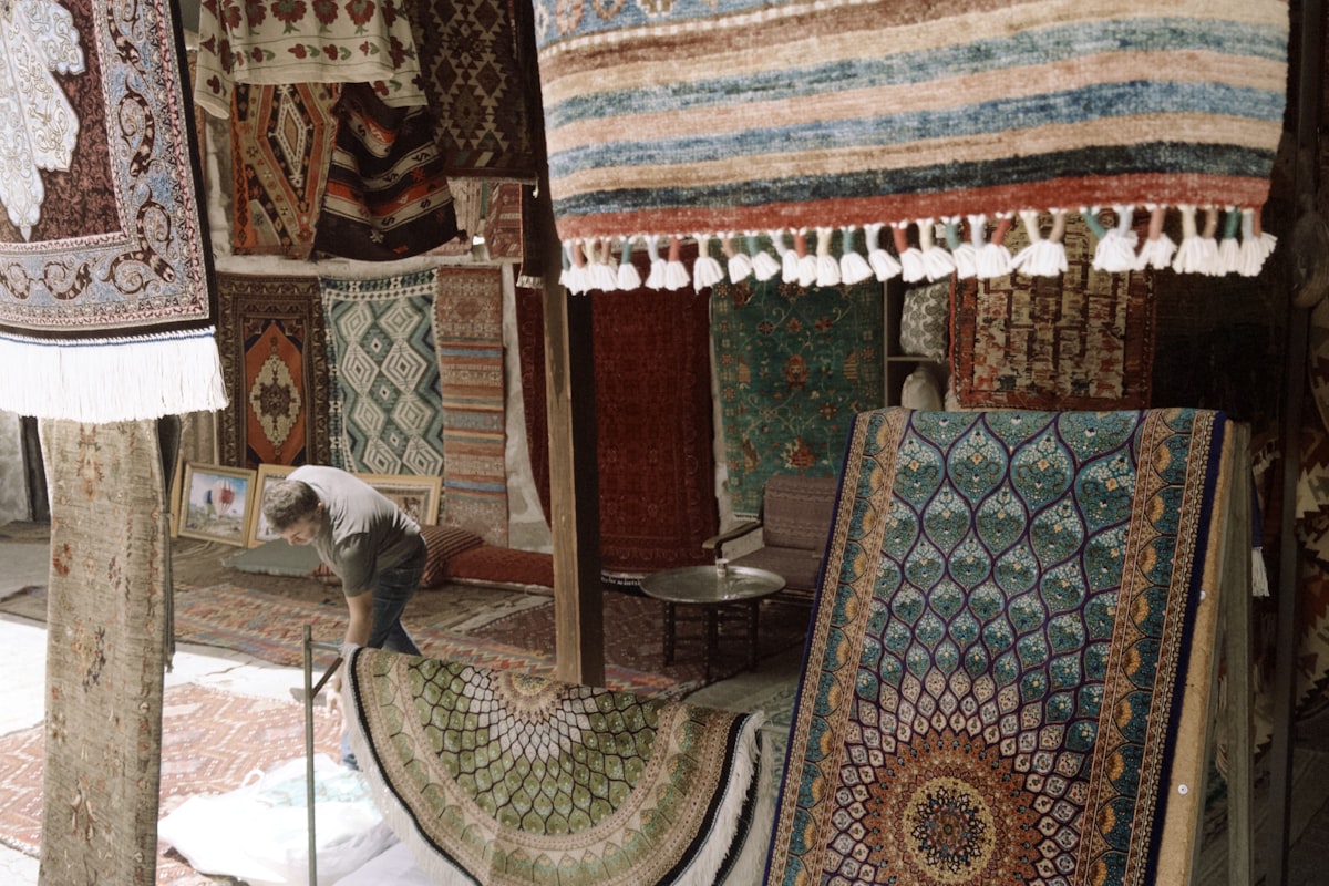 A person arranging patterned rugs in a shop filled with hanging textiles related to Explore Stylish Rugs for Every Home