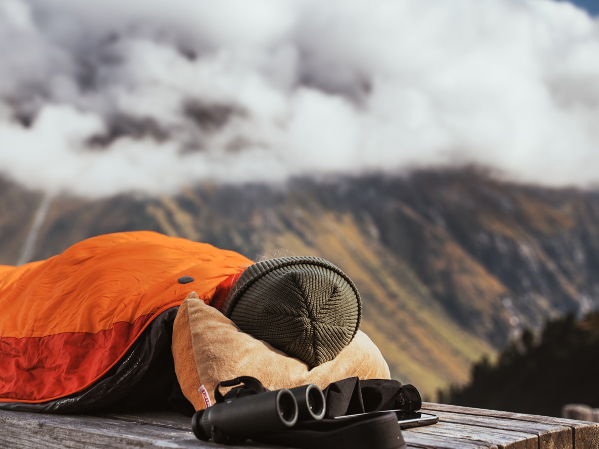 A person in an orange sleeping bag, lying on a wooden surface outdoors, likely in a mountainous area. Binoculars are nearby, and the foggy mountain backdrop creates a peaceful, remote, and reflective atmosphere. related to Warming Up to the Great Outdoors: The Ultimate Guide to Extra Large Cast Iron Chimineas