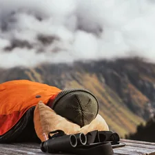 A person in an orange sleeping bag, lying on a wooden surface outdoors, likely in a mountainous area. Binoculars are nearby, and the foggy mountain backdrop creates a peaceful, remote, and reflective atmosphere. related to Warming Up to the Great Outdoors: The Ultimate Guide to Extra Large Cast Iron Chimineas