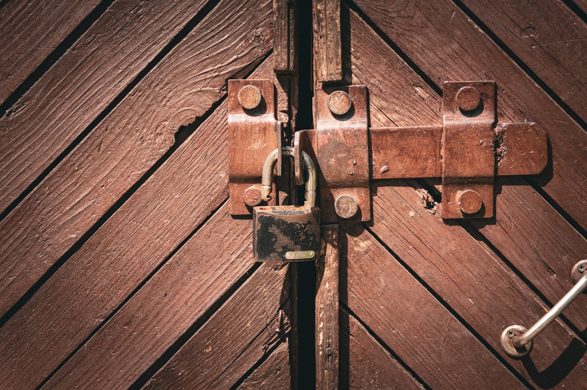 A rusty padlock and heavy metal bolt securing an old wooden gate, weathered brown planks laid in a chevron pattern. Warm, textural close-up of a locked barn or garage door. related to Unlock Your Entryway Potential: DIY Locker Plans for Every Home