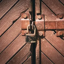 A rusty padlock and heavy metal bolt securing an old wooden gate, weathered brown planks laid in a chevron pattern. Warm, textural close-up of a locked barn or garage door. related to Unlock Your Entryway Potential: DIY Locker Plans for Every Home