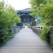 A scenic walkway on a metal bridge with wooden planks is framed by lush green foliage on either side. The bridge's black iron framework stands out against the verdant backdrop, with lampposts lining the path, creating a serene and inviting atmosphere. The path leads towards a distant, tree-covered hill. related to Discover the Charm of Missoula Bed and Breakfasts
