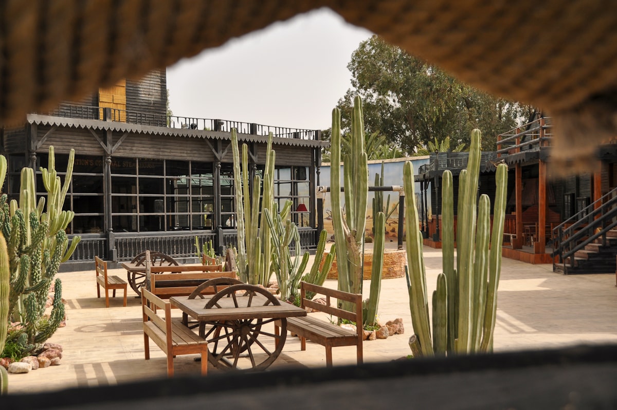 A western-themed courtyard framed by a rustic opening, featuring wooden tables with wagon wheel details, tall cacti, and classic saloon-style wooden buildings under a warm desert-like sky. 📸 Photo by me IG: @margoevardson related to Spice Up Your Outdoor Space with Chimineas on Sale