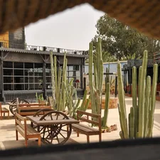 A western-themed courtyard framed by a rustic opening, featuring wooden tables with wagon wheel details, tall cacti, and classic saloon-style wooden buildings under a warm desert-like sky. 📸 Photo by me IG: @margoevardson related to Spice Up Your Outdoor Space with Chimineas on Sale
