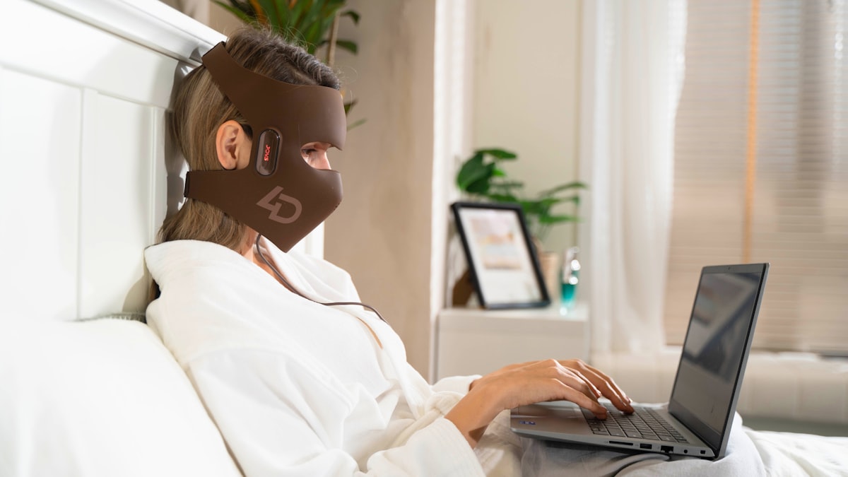 A woman in a white robe sits on her bed, using the JOVS 4D Laser Light Therapy Mask while tapping on her laptop. related to Brewing Up Comfort: Bed Bath Coffee Makers for the Perfect Morning
