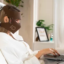 A woman in a white robe sits on her bed, using the JOVS 4D Laser Light Therapy Mask while tapping on her laptop. related to Brewing Up Comfort: Bed Bath Coffee Makers for the Perfect Morning