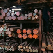 An industrial stockyard, featuring stacked hexagonal and circular metal bars on shelves. The varied ends of the bars display a spectrum of colors—from rust-red to oxidized green—creating a dense, repetitive pattern and geometric complexity. A worker on the right adds a sense of human scale to the heavy inventory. This texture is highly useful for engineering firm advertisements, as a conceptual background representing raw material wealth, or in editorial content about manufacturing and global supply chains. related to Discover the Elegance of Rod Iron Bed Frames