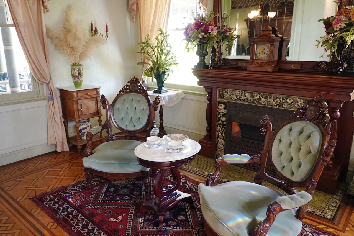 An inviting Victorian parlor at the Winchester Mystery House features carved wood armchairs with tufted velvet upholstery, a marble-topped table set for tea, and a richly tiled fireplace beneath a grand mantel. Floral arrangements and delicate furnishings add to the refined yet cozy charm. Sunlight and soft pastels give the room a warm, timeless elegance. related to Experience Elegance at a Grand Victorian Bed and Breakfast