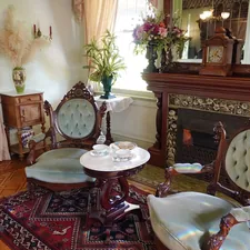 An inviting Victorian parlor at the Winchester Mystery House features carved wood armchairs with tufted velvet upholstery, a marble-topped table set for tea, and a richly tiled fireplace beneath a grand mantel. Floral arrangements and delicate furnishings add to the refined yet cozy charm. Sunlight and soft pastels give the room a warm, timeless elegance. related to Experience Elegance at a Grand Victorian Bed and Breakfast