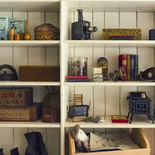 Antique object Collection Displayed On White Shelves - The image shows a collection of vintage toys and household items displayed on white wooden shelves. The collection includes an antique projector, a toy stove, a miniature bed with bedding, and various boxes of old-fashioned products. related to The Ultimate Guide to White Wooden Beds
