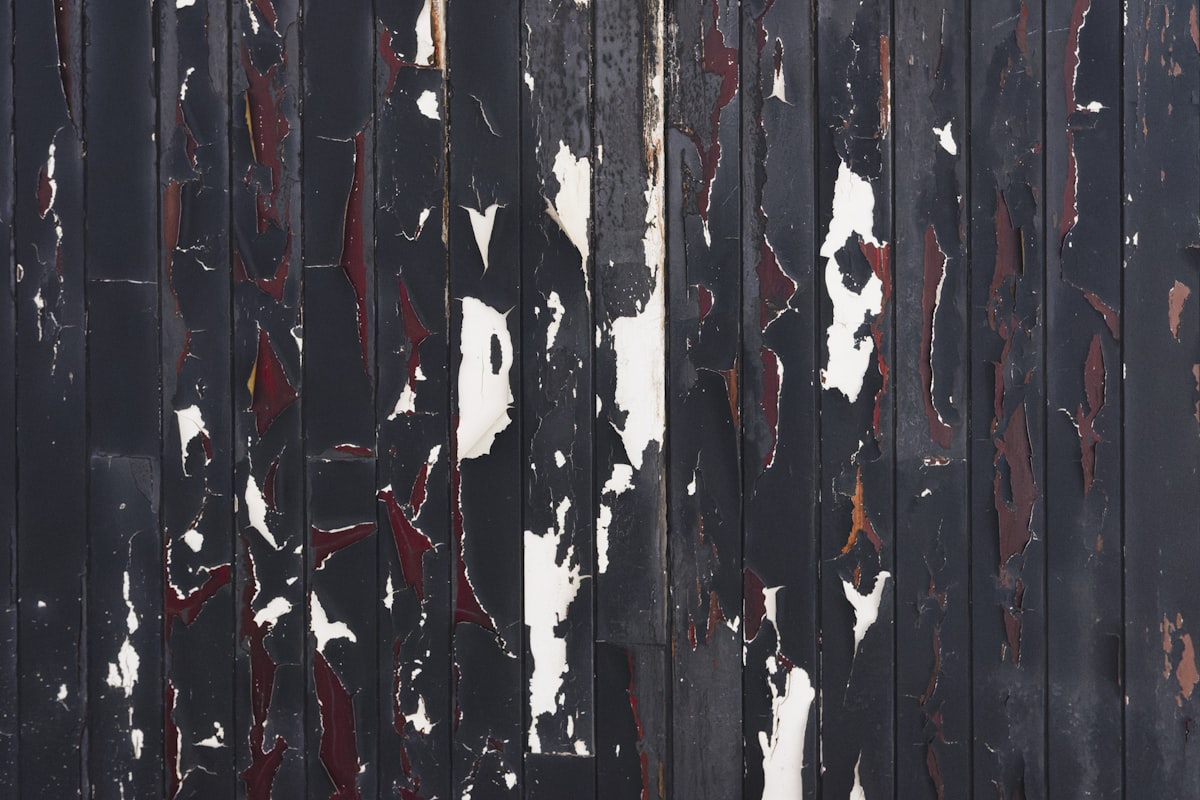 Close Up Of Peeling Black Paint On Wooden Paneling - A close up view of weathered black wooden paneling. The paint is peeling and chipping, revealing layers of red and white paint beneath. The texture is rough and uneven, creating an abstract pattern. related to Elevate Your Home Decor with a Black Distressed Sideboard