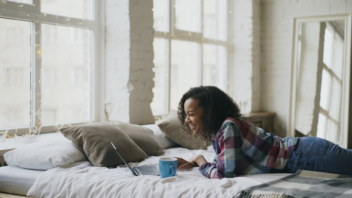 Curly mixed race student girl lauging while using laptop for sharing social media lying in bed at home related to Elevate Your Space with Bed Risers from IKEA