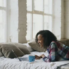 Curly mixed race student girl lauging while using laptop for sharing social media lying in bed at home related to Elevate Your Space with Bed Risers from IKEA