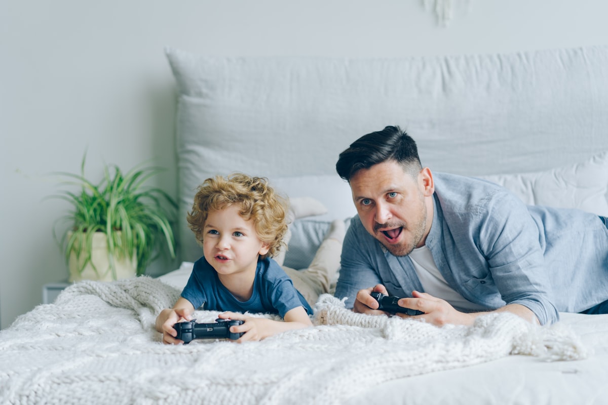 Father and child playing video game on bed at home pressing buttons on joystick related to Valances for Boys' Bedroom
