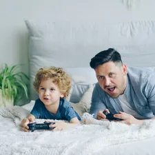 Father and child playing video game on bed at home pressing buttons on joystick related to Valances for Boys' Bedroom