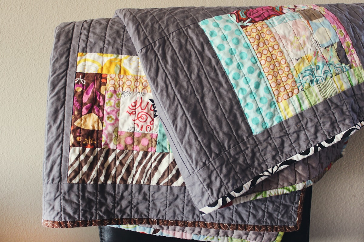 Handmade colorful quilt hangs over a chair, in colors of gray, blue, pink, brown and yellow with abstract designed quilt blocks. related to Purple Quilts & Coverlets: Warmth & Beauty in Every Shade