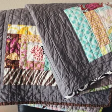 Handmade colorful quilt hangs over a chair, in colors of gray, blue, pink, brown and yellow with abstract designed quilt blocks. related to Purple Quilts & Coverlets: Warmth & Beauty in Every Shade