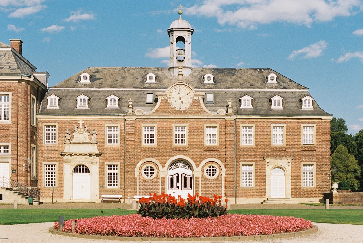 Historic palace facade symmetry analog photography architecture. Elegant European baroque-style building with red brick walls, central clock and flower garden, captured on Kodak Portra 800 film using a Contax camera related to Beautiful Flower Bed Designs for the Front of Your House