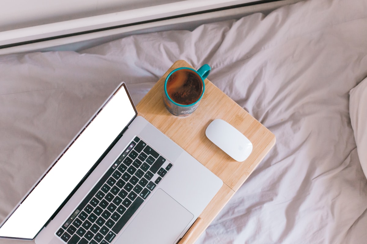 macbook pro beside white ceramic mug on white textile related to Elevate Your Sleeping Experience with Computer Tables for Bed