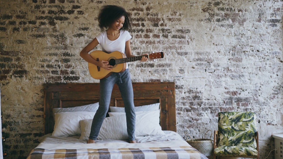 Mixed race young funny girl playing acoustic guitar and have fun dancing on bed at home on holidays related to Elevate Your Teen's Bedroom Style with PB Teen Bedding