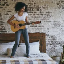 Mixed race young funny girl playing acoustic guitar and have fun dancing on bed at home on holidays related to Elevate Your Teen's Bedroom Style with PB Teen Bedding