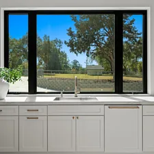 Modern kitchen sink with white shaker cabinetry, brass hardware, and large black-framed picture window overlooking greenery. Bright contemporary home interior featuring marble countertops, natural light, and clean minimalist design. High-end residential kitchen with timeless finishes and warm wood accents. related to Kitchen Window Valance Ideas to Elevate Your Cooking Space