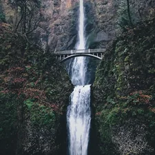 Moody photo of a bridge over a stunning double waterfall between two lush green mountains with lots of moss, leaves, ferns, and evergreen trees. related to Discover the Charm of Hood River Bed and Breakfasts