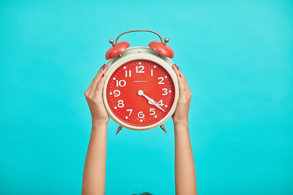 person holding red and beige twin bell analog alarm clock related to The Sweet Spot: What Time Do I Need to Go to Bed?