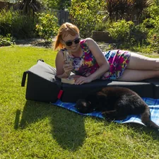 Smiling woman wearing sunglasses in a short, colourful summer dress lying in the sun on a padded lounger and a blue picnic blanket in the garden holding a glass of champagne with her cat lying next to her related to Choosing the Perfect Dog Ramp for Your Bed: A Comprehensive Guide