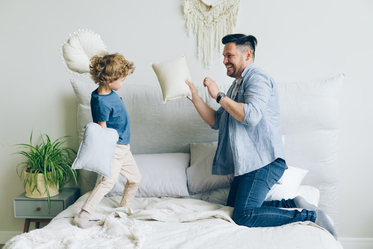 Young man fighting pillows with little son having fun on bed in apartment related to Elevate Your Bedding Game with Quilted Coverlets and Shams