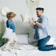 Young man fighting pillows with little son having fun on bed in apartment related to Elevate Your Bedding Game with Quilted Coverlets and Shams